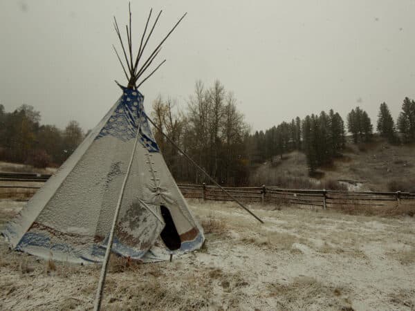 A snow-dusted tipi stands in a grassy field on a ranch for sale, surrounded by a wooden fence, leafless trees, and pine-covered hills. Snowflakes gently fall on this wintry day.