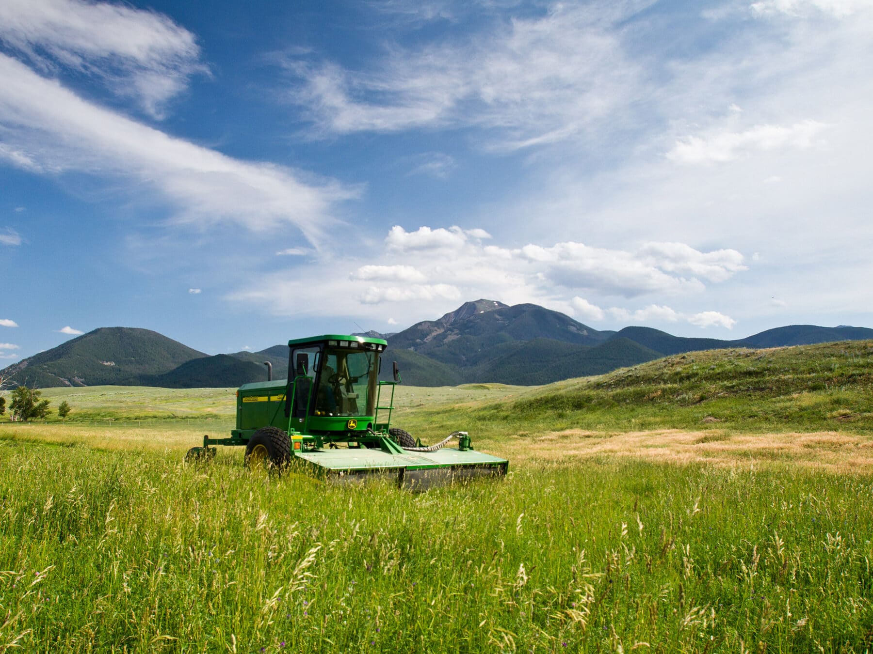 A green tractor sits in a tall, grassy field under a blue sky with scattered clouds, with rolling hills and mountains in the background—perfect scenery for a ranch for sale or recreational land.
