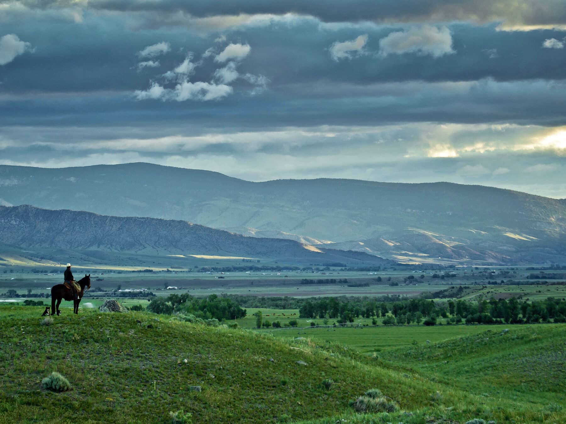 A lone person on horseback stands on a grassy hill, overlooking a wide valley ideal for a hunting property, with scattered trees and distant mountains under a dramatic, cloudy sky.