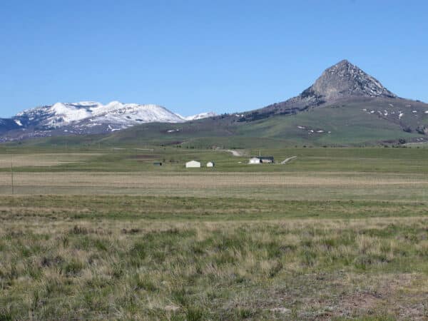A grassy plain with scattered buildings sits beneath a pyramid-shaped mountain; snowy peaks rise in the background under a clear blue sky, offering an ideal cattle ranch or land for sale.