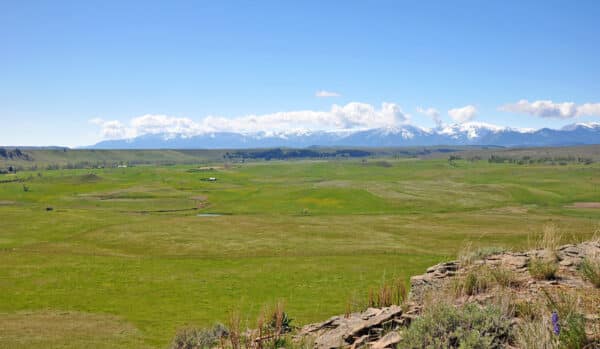 Vast green fields stretch under a clear blue sky, with snow-capped mountains in the distance. Scattered buildings and ponds dot this scenic cattle ranch for sale, with some rocky terrain visible in the foreground.