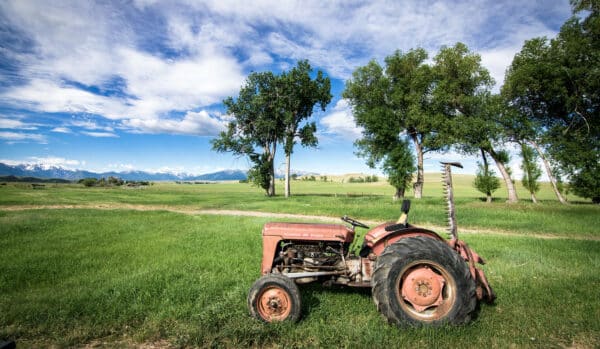An old red tractor sits on green grass in a rural field, perfect as a ranch for sale, with tall trees nearby and distant snow-capped mountains under a partly cloudy blue sky.