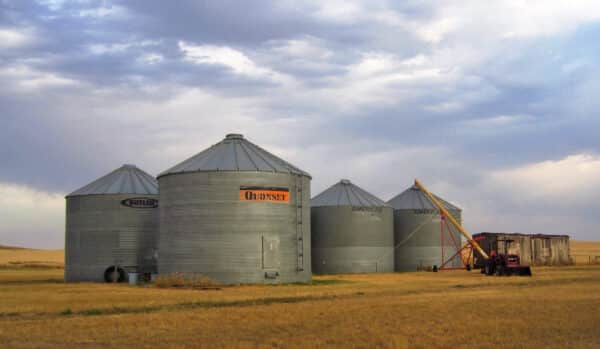 Four metal grain silos stand on a grassy field under a cloudy sky. A red tractor with a trailer and auger is parked beside the silos, highlighting the potential of this recreational land or hunting property.