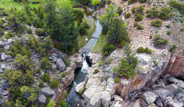Aerial view of a small waterfall flowing through rocky terrain surrounded by green trees and shrubs, with a winding river continuing through the landscape—ideal setting for land for sale or a scenic cattle ranch.