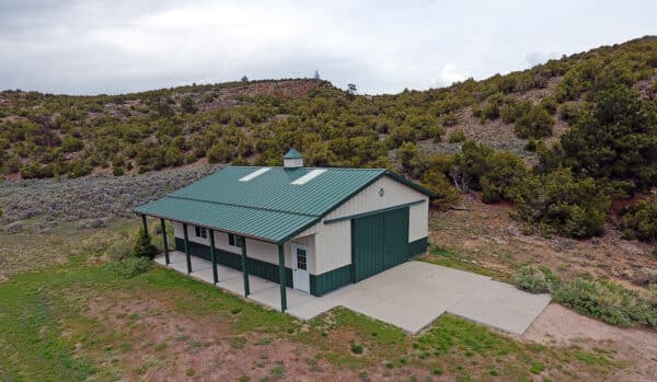 A small green and white metal building with a covered porch stands on a concrete pad, surrounded by grass and shrubs at the base of a tree-covered hillside—ideal recreational land or potential ranch for sale.