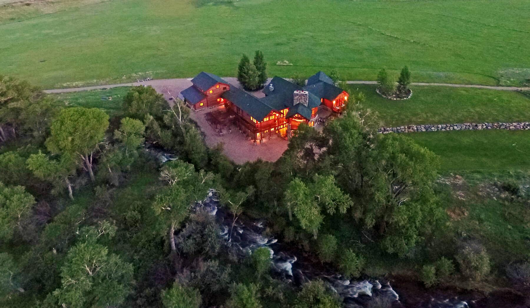 Aerial view of a ranch for sale, a large, warmly lit house surrounded by trees and near a river, set in a green, open landscape during dusk.