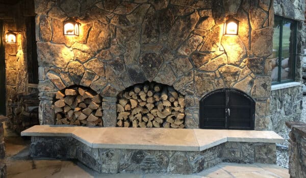 A rustic stone outdoor fireplace on recreational land features two arched wood storage spaces filled with firewood, a black metal oven door, a stone bench, and two wall-mounted lanterns casting a warm glow.