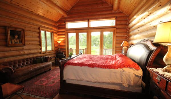 A cozy log cabin bedroom with a wooden bed, red blanket, rustic furniture, brown leather sofa, red rug, lamps, and artwork. Large windows fill the space with natural light and offer lush views—perfect for your dream cattle ranch for sale.