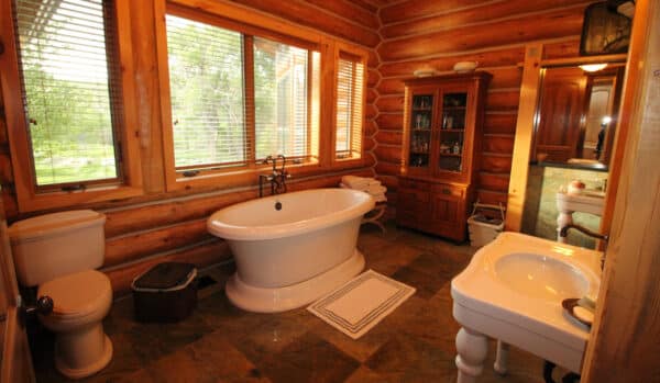 Log cabin-style bathroom on a cattle ranch with wood walls, freestanding white bathtub, toilet, white sink, wooden cabinet, large windows with blinds, and green-tiled floor. Towels are stacked on a shelf beside the tub.