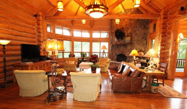 A spacious log cabin living room with wood walls and ceiling, large windows, cozy armchairs, a leather sofa, and a stone fireplace. Warm lighting and rustic decor create an inviting atmosphere on this exceptional ranch for sale.