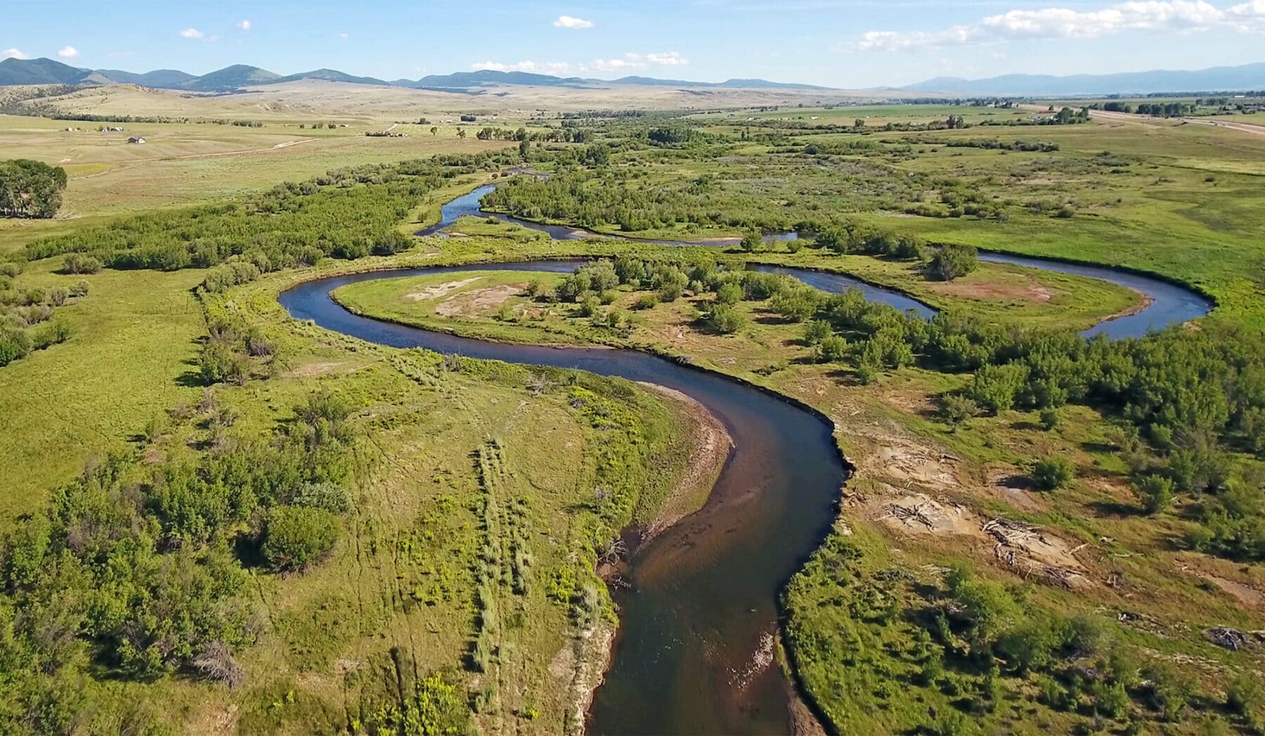A winding river curves through a lush green landscape with fields and shrubs, ideal for a cattle ranch, backed by distant mountains under a partly cloudy blue sky.