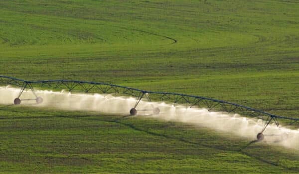 A center pivot irrigation system waters a large, green agricultural field—ideal for a cattle ranch or hunting property—spraying fine mist in a circular pattern as it moves across the landscape.