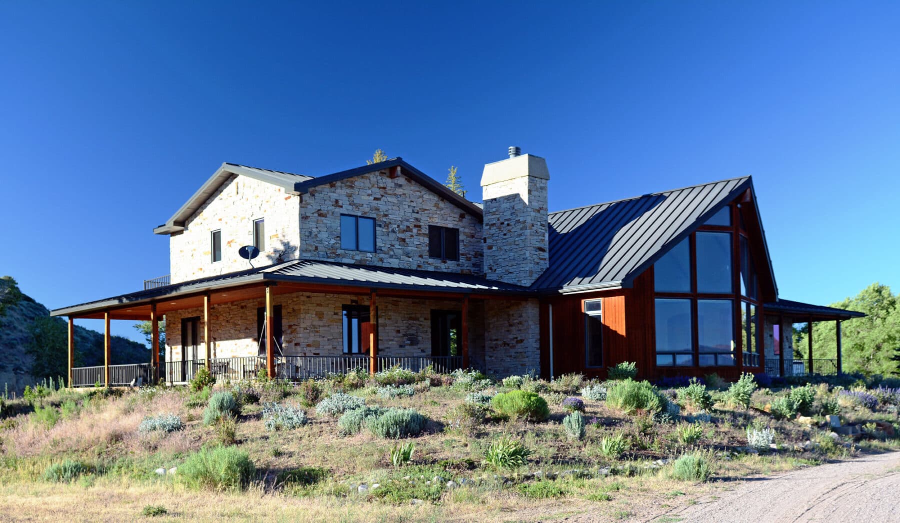 A modern two-story house with stone and wood exterior, large windows, a black metal roof, and a wraparound porch sits on a grassy plot of recreational land under a clear blue sky.