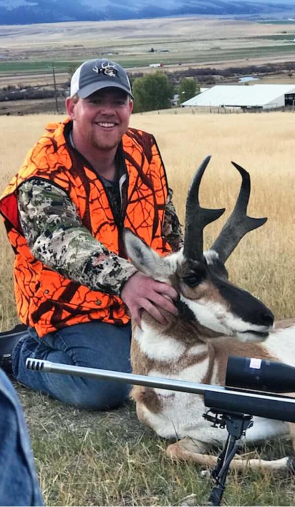 A smiling hunter in an orange camo vest kneels on recreational land, holding a pronghorn antelope by the horns. A rifle lies in front of him, with open fields and buildings in the background.