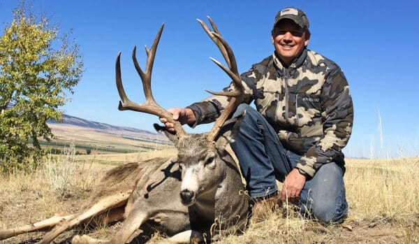 A person in camouflage clothing kneels on dry grass next to a large deer with antlers, resting one hand on the deer. Mountains and blue sky fill the background—ideal scenery for recreational land or ranch for sale.