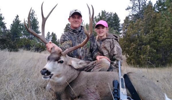 Two people in camouflage, one adult and one child, pose outdoors with a large deer they have hunted on a hunting property. The adult holds the deer's antlers while a rifle rests nearby. Trees form the background.