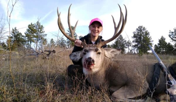 A person in a pink cap smiles while kneeling beside a large deer with antlers on recreational land in a grassy, lightly wooded area. A rifle lies next to the deer, highlighting the appeal of quality hunting property.