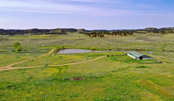 Aerial view of a rural landscape featuring green fields, a small pond, dirt roads, scattered trees, and a white barn with a green roof under a blue sky—ideal cattle ranch or recreational land for sale.