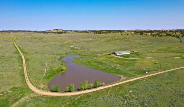 Aerial view of a rural landscape with a dirt road curving past a pond surrounded by greenery, near a large barn and open fields—perfect recreational land or hunting property under a clear blue sky.
