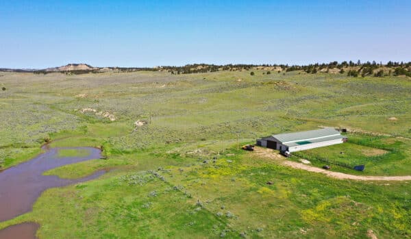 A large metal barn sits on a grassy, rolling plain near a dirt road and pond, perfect for a cattle ranch or recreational land. Distant hills and sparse trees stretch under a clear blue sky, highlighting this unique land for sale.