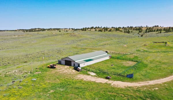 A large green-roofed barn sits in a fenced area on open grassy land for sale with rolling hills, a dirt road, truck, and scattered trees under a clear blue sky—ideal as a hunting property or cattle ranch.