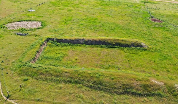 A rectangular, shallow, grassy depression in a green open field—ideal for a cattle ranch or hunting property—bordered by taller grass, with two circular patches of dirt nearby. Fencing and sparse vegetation are visible in the background.