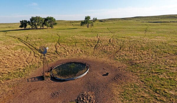 A round water trough with a windmill stands on dry, open grassland—classic features of a cattle ranch. Trails from livestock lead to the trough, with scattered trees and rolling hills in the distance under a blue sky. Ranch for sale.