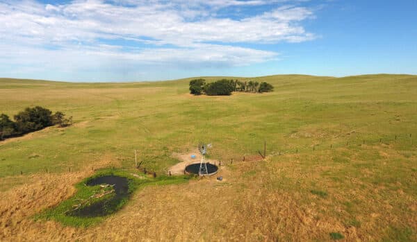 Aerial view of a grassy field with a small pond, fenced-in area, and scattered trees under a partly cloudy sky—ideal as a hunting property or cattle ranch. Ranch for sale.