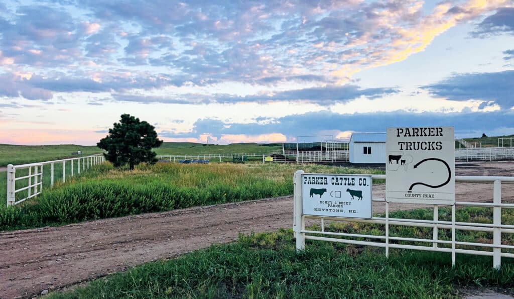 A rural cattle ranch with grassy fields, a dirt road, fenced enclosures, and a white sign reading Parker Cattle Co. and Parker Trucks under a partly cloudy sunset sky—ideal recreational land or hunting property.