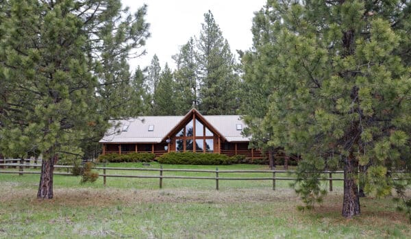 A log cabin with large windows is nestled among tall pine trees, surrounded by grass and a wooden fence, making it an ideal hunting property with a scenic forested background.