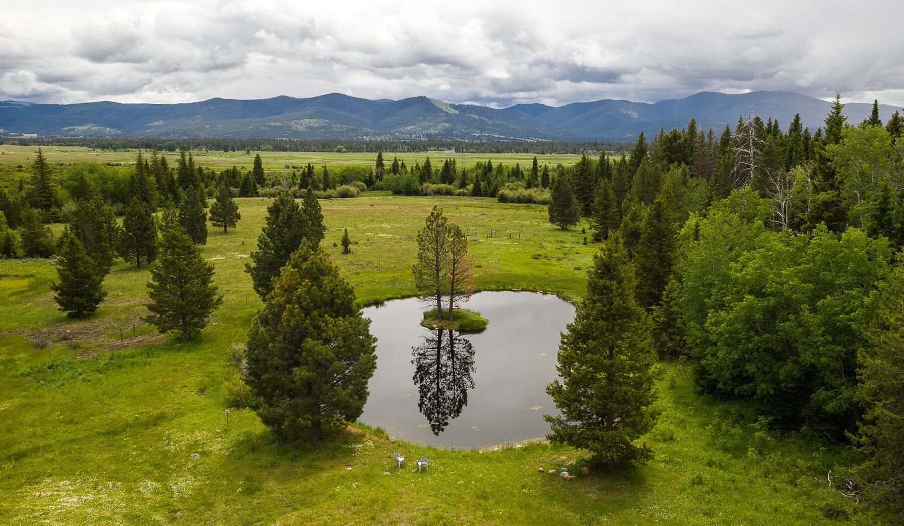 A small, circular pond surrounded by green trees and grassy fields highlights this picturesque land for sale, with a single tree reflected in the water. Mountains and a cloudy sky create a serene backdrop for your future cattle ranch.