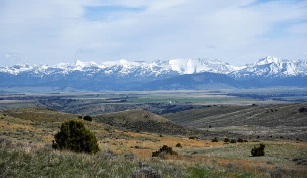 Rolling grassy hills with scattered shrubs in the foreground, farmland and cattle ranch in the midground, and a distant range of snow-capped mountains under a partly cloudy sky.