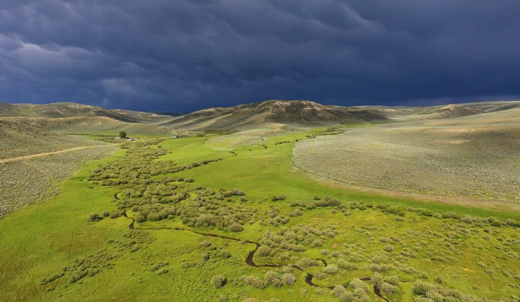 A winding creek flows through a lush green valley surrounded by rolling hills under a dramatic, stormy sky. A small house sits in the distance, highlighting this recreational land for sale amid the expansive landscape.