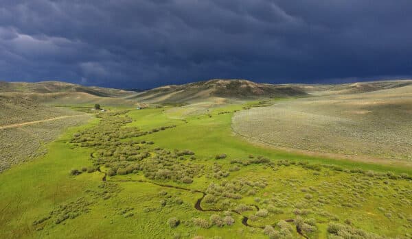 A winding creek flows through a lush green valley surrounded by rolling hills under a dramatic, stormy sky. A small house sits in the distance, highlighting this recreational land for sale amid the expansive landscape.