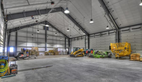 Large, clean warehouse with tall ceilings and bright lighting—perfect for a working cattle ranch or recreational land needs. Snowmobile, skid steer, green mower, and yellow wood chipper are parked along the right wall; pallets and supplies in back.