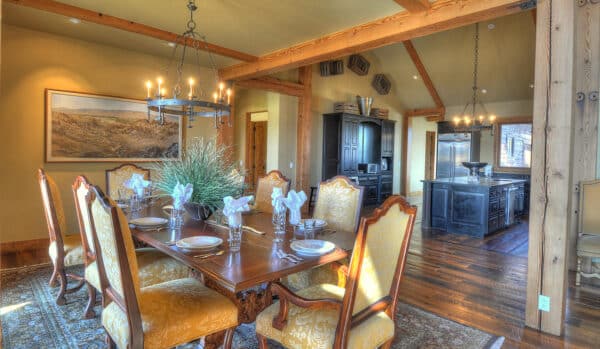 Elegant dining room with a wooden table set for six, ornate chairs, and a chandelier overhead. In the background of this ranch for sale, a modern kitchen with dark cabinets and stainless steel appliances is visible. Warm lighting and wooden beams create a cozy atmosphere.