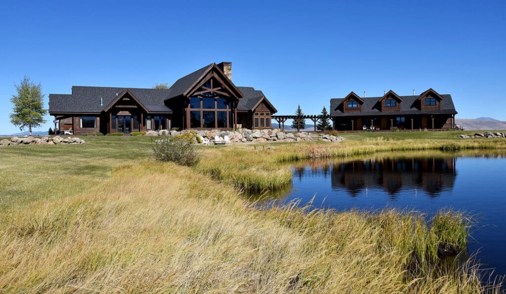 A large rustic house and detached guesthouse sit on a grassy lawn by a pond, with clear blue sky and distant mountains. Ideal as a hunting property or cattle ranch, the picturesque scene is reflected in the calm water.