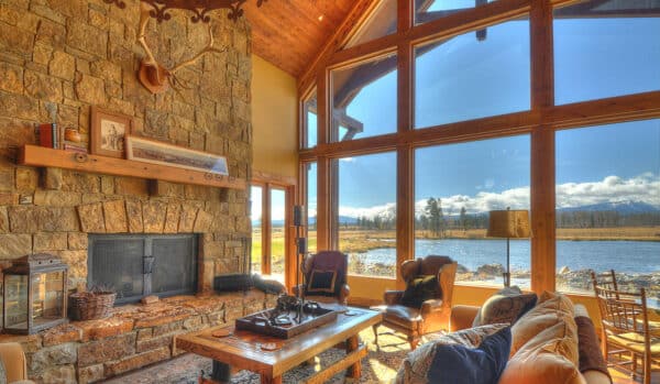 A cozy living room with stone fireplace, antler decor, and large windows reveals a scenic view of a river and mountains under a bright blue sky—perfect for relaxing on this hunting property, where rustic wooden furniture and warm sunlight fill the space.
