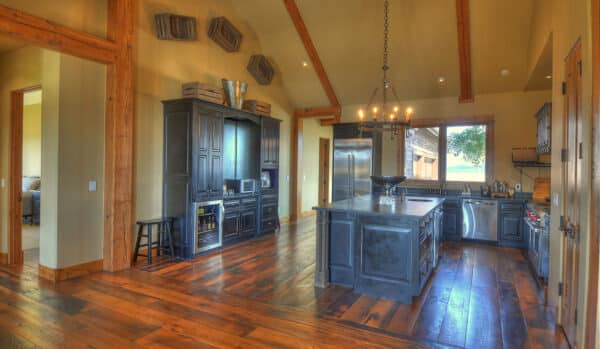 Spacious rustic kitchen with dark cabinets, wooden beams, and wide-plank wood floors—perfect for a cattle ranch. An island with a sink sits in the center beneath a chandelier. Large windows bring in ample natural light.