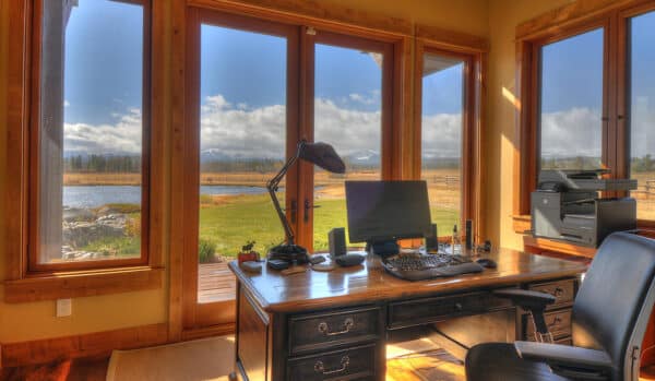 A home office with a wooden desk, computer, lamp, and printer sits by large windows overlooking recreational land for sale with a scenic lake, grassy field, and distant mountains under a partly cloudy sky.