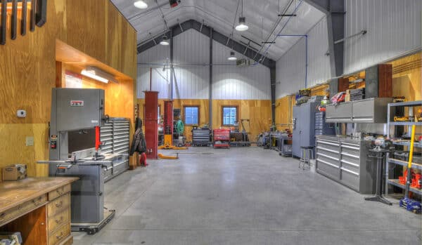 Spacious, well-lit workshop with high ceilings, organized tool cabinets, workbenches, and various industrial machines along the walls—ideal for maintaining equipment on a hunting property or cattle ranch. Concrete floors and wood-paneled sections add warmth to the space.