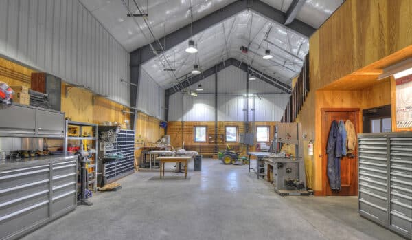 Spacious, well-lit workshop with high ceilings, metal cabinets, workbenches, tools, and wooden walls—ideal for a hunting property or as part of a land for sale. Features a loft area with stairs and organized storage along the sides.