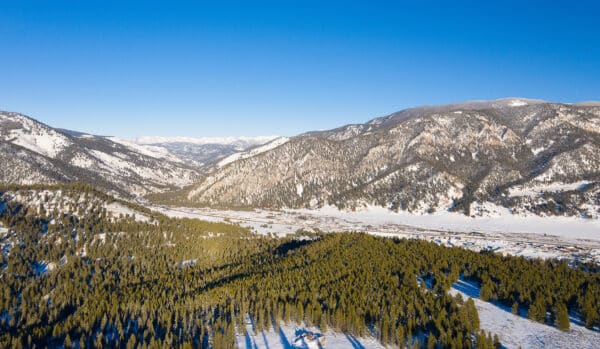 Aerial view of a snow-covered mountain valley with dense evergreen forests, a clear blue sky, and a small town nestled between the mountains—ideal recreational land or hunting property for those seeking adventure.