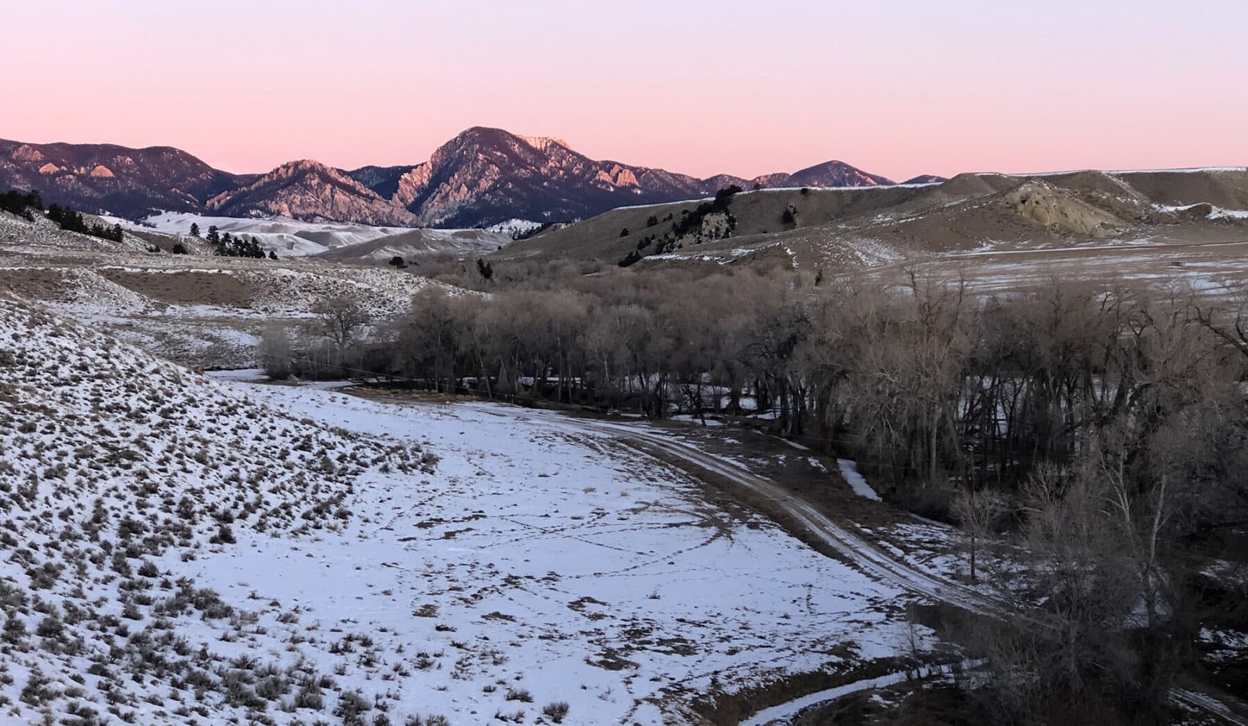Snow-dusted hills and bare trees in a wide valley, with rugged mountains in the background illuminated by pink sunset light—a stunning hunting property or ranch for sale under a clear sky.