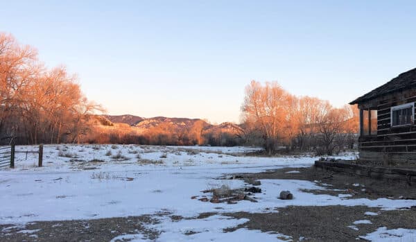 Snow-covered ground with patches of grass and bare trees lit by golden sunlight. Mountains rise in the distance under a clear sky, and a rustic wooden cabin sits on the right side—ideal hunting property or ranch for sale.