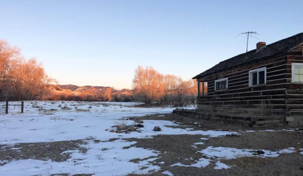A rustic log cabin stands on recreational land amid a snowy, barren landscape at sunset, with leafless trees and distant hills bathed in warm orange light under a clear blue sky.