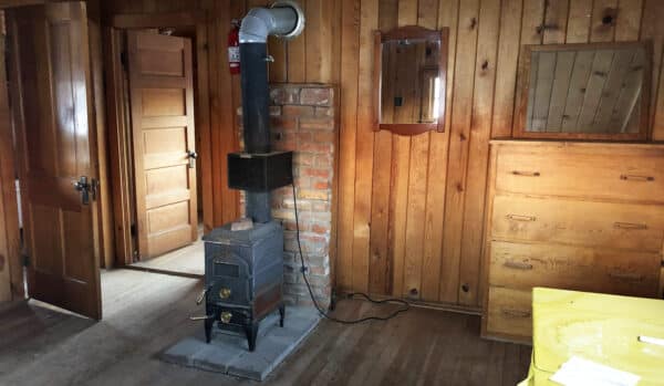 A small wood-burning stove sits on a stone base against a brick and wood-paneled wall in a rustic room, featuring wooden doors, a dresser, yellow countertop, and mirror—perfect for your dream ranch for sale or hunting property.