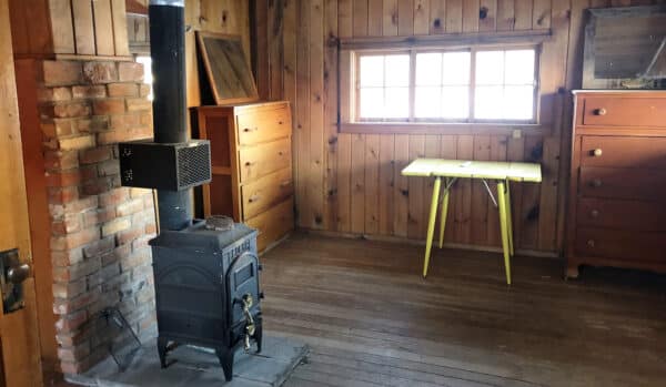 A rustic wooden room on a cattle ranch with a wood-burning stove near a brick wall, a small yellow table by the window, and several wooden dressers. Sunlight streams in, illuminating this inviting ranch for sale.