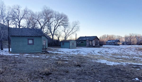 Several small, weathered green and brown houses stand on a snowy, patchy field with leafless trees in the background under a clear sky. This peaceful hunting property offers unique recreational land for outdoor enthusiasts.