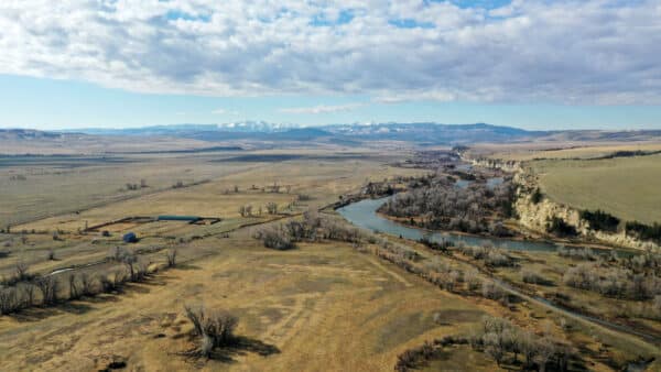 A wide river winds through an open plain with sparse trees and distant mountains under a partly cloudy sky. Farmland, a small structure, and a potential cattle ranch or hunting property border the scenic riverbank.
